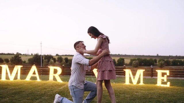 Young man asking for his girlfriend in marriage while sitting on one knee. Romantic evening request. Concept of marriage applications in nature. Merry me out of the lights. He said yes.
