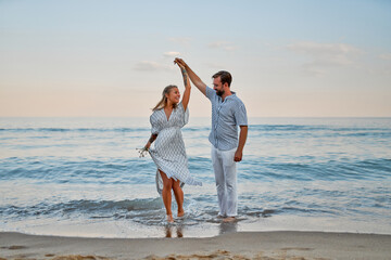 An attractive woman in a dress and a handsome bearded man in a shirt and white trousers are dancing in love on the seashore, having fun on vacation.