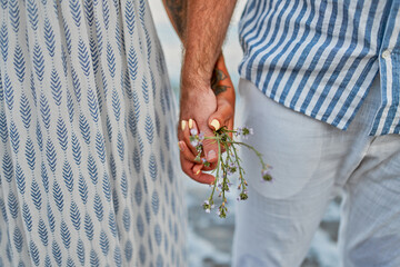 Cropped close-up of a couple holding hands with wildflowers in front of the sea.