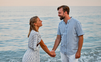 An attractive woman in a dress and a handsome bearded man in a striped shirt hold hands and romantically spend time on the seashore. A loving couple on vacation.