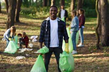 Stylish bearded black man with full rubbish packs on background of his friends activists picking up...