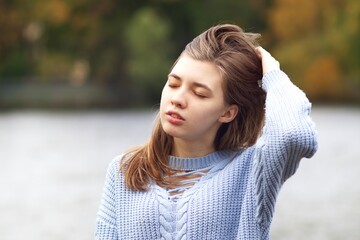 Portrait of young beautiful woman at autumn park with lake standing with eyes closed, enjoying good weather, breath deeply fresh air, relaxing