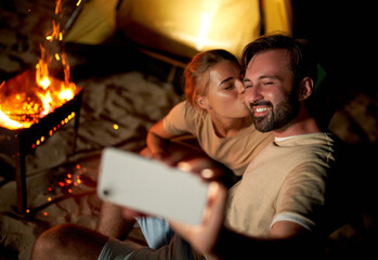A cute woman and a handsome man romantically spend time near the tent by the fire, take a selfie on a smartphone at night on the beach by the sea.