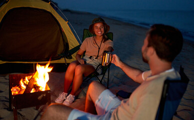 A young couple in love are sitting near the tent on folding chairs by the fire, drinking tea at night on the seashore.
