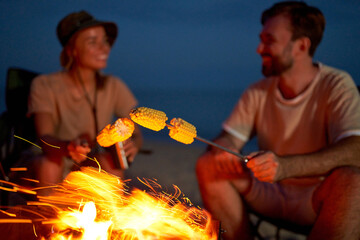 Young attractive couple sit on folding chairs near the tent and grill corn on a fire and have fun talking at night near the sea.