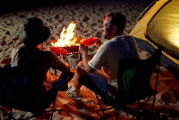 A cute woman and a handsome man are sitting on folding chairs near the tent by the fire, eating watermelon and having fun at night on the beach by the sea.
