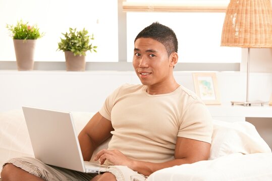 Young asian man sitting on sofa at home browsing Internet working in home office on laptop, smiling.