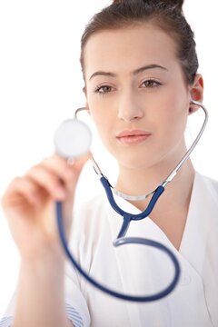Young woman in lab coat holding stethoscope towards camera, looking serious. Isolated on white.