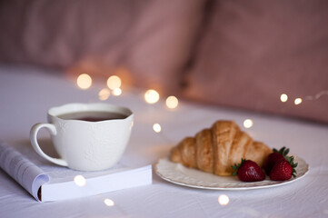 Cup of coffee on open paper book with fresh croissant in bed over Christmas lights close up. Good morning. Breakfast.
