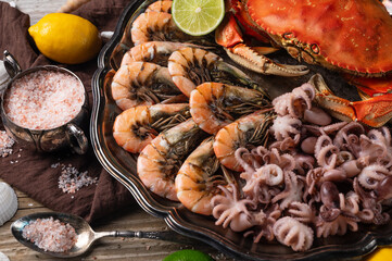 Close-up view of large dish with coked crab, baby octopuses and tiger shrimps served with lemon, lime, seashells and bowl with sea salt on rustic wooden background. Sea delicious.