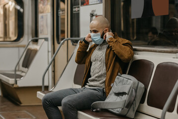 A man with a beard is putting on a medical mask on his face to avoid the spread of coronavirus in a subway car. A bald fellow in a surgical face mask against COVID-19 is sitting on a metro train.