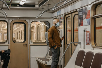 A man with a beard in a face mask to avoid the spread of coronavirus is waiting for a new stop in a subway car. A bald guy in a surgical mask against COVID-19 is holding a cellphone on a metro train.