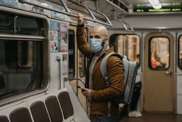 A man with a beard in a medical face mask to avoid the spread of coronavirus is looking around in a subway car. A bald guy in a surgical mask against COVID-19 is holding a cellphone on a metro train.