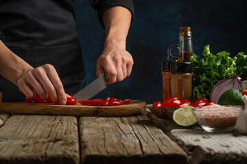 Close-up view of the chef cuts with knife cherry tomatoes on wooden chopped board on dark blue background. For the preparation of pizza, tomato sauce, salad. A delicious meal concept.