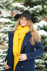 Outdoors lifestyle portrait of beautiful blonde girl smiling and walking in the snowy park. Wearing stylish dark blue coat, black hat, and mustard scarf. Bright colors. Positive emotions.
