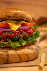 Close-up view of big tasty burger with beef, tomato, lettuce, red onion, cheese, mustard and ketchup on wooden chopped board with french potato. Concept of unhealthy street food. Appetizing meal.