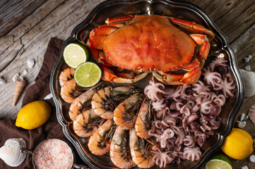 Top view of big dish with cooked crab, baby octopuses and tiger shrimps served with sea salt, lemon, lime and white seashells on rustic wooden background. Seafood concept. Delicious food.