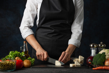 The professional chef in black apron cuts with knife cheese feta on black chopped board for cooking...