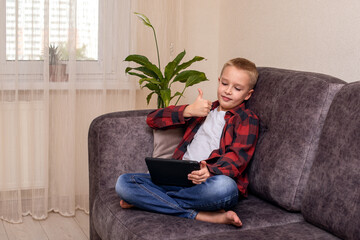 A charming 10-year-old boy in jeans and a checkered red shirt, wearing glasses and large headphones is listening to music or listening to a lesson on his smartphone