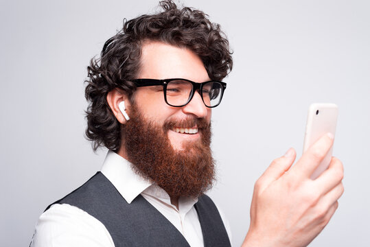 Close up portrait of young bearded hipster man using smartphone and earpods over white wall