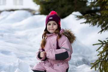 A cute little girl sits in the snow near green fir trees. Ski resort. A child in overalls against the background of a winter landscape. Winter walks, fun outdoors winter time.