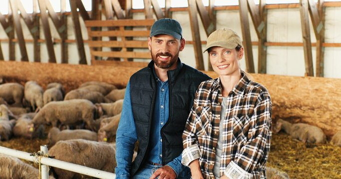 Portrait of happy young Caucasian married couple of farmers looking at each other and smiling to camera while sitting in stable for livestock. Handsome man and beautiful woman at sheep farm.