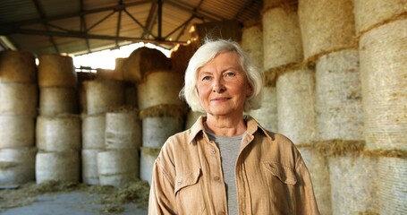 Portrait of Caucasian old gray-haired woman shepherd standing in barn with hay stocks on background...