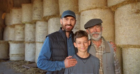 Three male generations in shed at hay stocks. Caucasian grandfather, father and son smiling to camera and standing in barn. Farming concept. Countryside. Old man farmer with son and grandson.