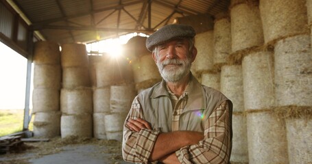 Portrait of Caucasian old gray-haired male farmer in cap standing at hay stocks in farm and looking at camera in sunlight. Grandfather worker at village. Countryside concept. Senior man. © VAKSMANV
