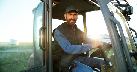 Porträt eines jungen kaukasischen männlichen Bauern mit Mütze, der mit offener Tür im Traktor sitzt und in die Kamera lächelt. Ackerbaufahrzeug. Maschine für die Landwirtschaft. Schöner lächelnder Mann.