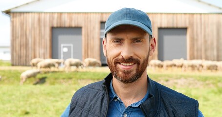 Portrait of Caucasian young smiled happy man with beard posing to camera with hands on sides and smiling. Handsome male farmer standing at field pasture with sheep grazing on background.