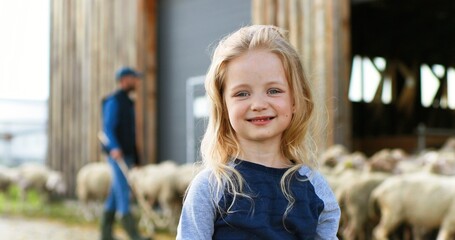 Portrait Caucasian small girl standing outdoor at livestock farm and smiling to camera. Sheep flock and man shepherd on background. Little cheerful kid laughing while spending summer at wool farming.