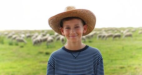 Portrait of Caucasian teen small happy boy in hat standing in green field in summer and smiling to camera. Sheep flock grazing on background. Little teenager shepherd. Vacations at countryside farm.
