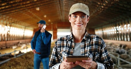 Young Caucasian woman using tablet device and working in farm stable. Female farmer tapping and scrolling on gadget computer in shed. Man talking on phone on background. Male speaking on cellphone.