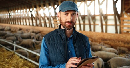 Portrait of Caucasian man shepherd smiling at camera in shed with cattle animals and using tablet...