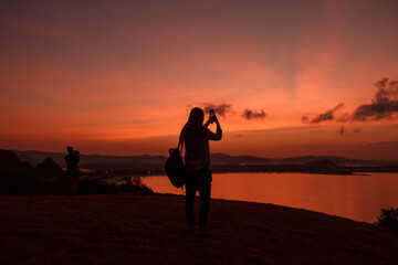 Fototapeta premium Silhouette woman take picture sunset by the sea on mobile phone. Tourist with backpack photographing ocean red sunset sky