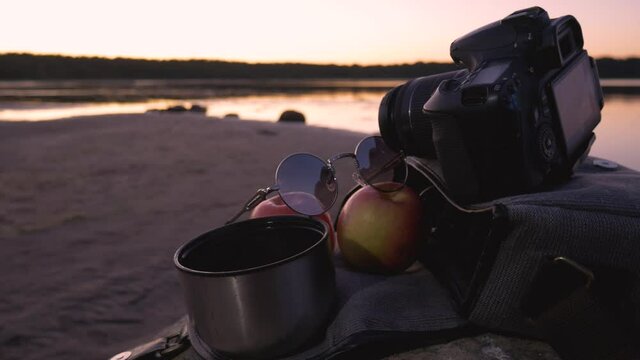 Photographer's bag with 2 apples and coffee. Tourist halt on the stone against the background of the lake and a beautiful sunset.
