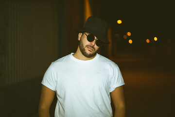 Chico joven trapero vistiendo ropa streetwear urbano con flash con gafas en un callejon en el sur...