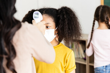 A group of Kids students wearing masks lined up waiting for woman teachers to Check Fever by Digital Thermometer in the classroom for Scan and Protect from Coronavirus Outbreak - Healthcare Concept