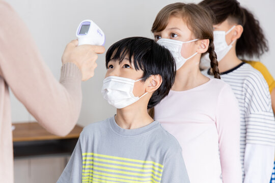 A group of Kids students wearing masks lined up waiting for woman teachers to Check Fever by Digital Thermometer in the classroom for Scan and Protect from Coronavirus Outbreak - Healthcare Concept