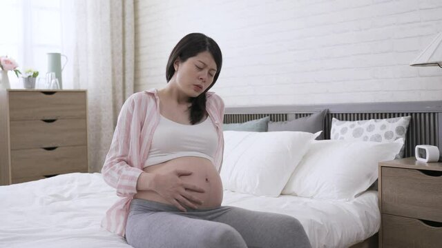 Korean expectant mom sitting alone on bed is moaning and stroking her abdomen while suffering painful contraction at background bright home interior.