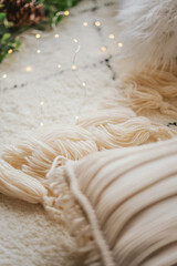 Knitted beige plaid with tassels and macrame pillow on a white fluffy carpet. Christmas comfort in the interior.