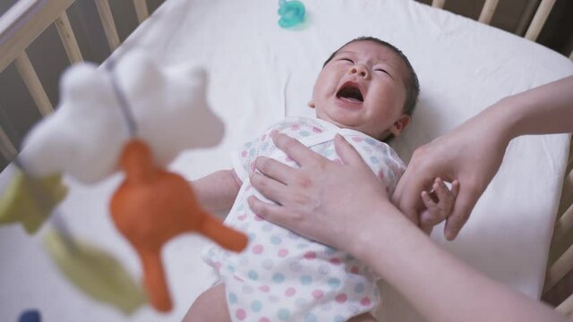 chinese mother is soothing her restless and crying newborn child by gently touching her on the chest and head in the crib with a colorful mobile.
