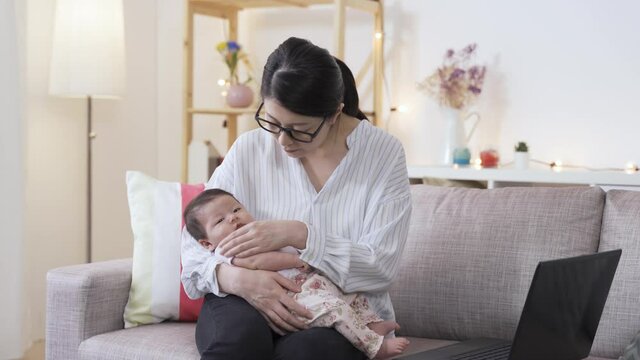 Portrait Asian Multitasking Mom Is Coaxing Her Lovely Baby In Her Arms While Working On The Laptop In The Living Room At Home.