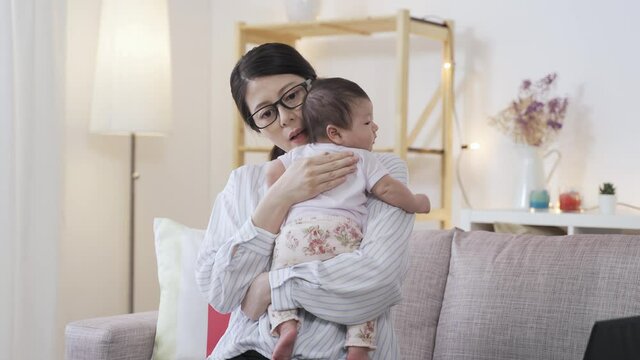 portrait chinese working mother is burping her adorable baby by patting on its back on the sofa in a cozy living room.
