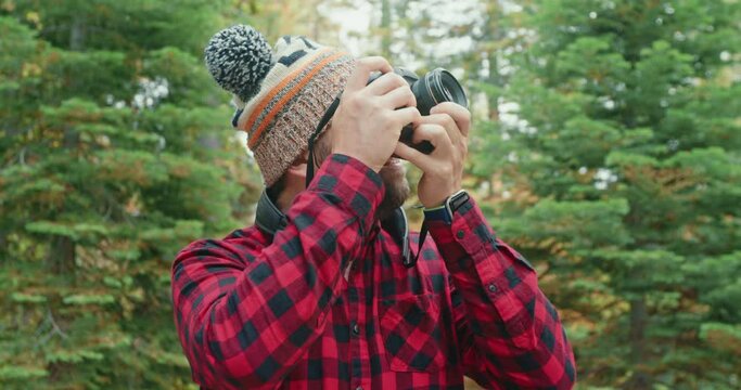 Adult bearded man in hat with a camera taking pictures of early fall nature on a background of green forest with pine trees. Smiling guy photographing scenic woods. Outdoor adventure and nature travel