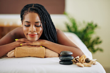 Smiling young Black woman lying on bed in spa salon with eyes closed after getting rejuvenating treatment