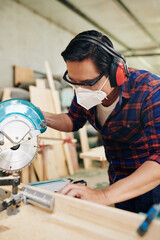 Carpenter in earmuffs, mas and goggles cutting wooden plank with circular saw