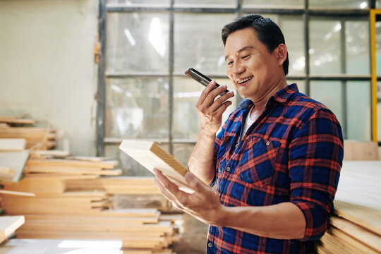 Smiling Asian Carpenter Looking At Wooden Brick In His Hand When Recording Voice Message To Customer