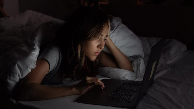 Young Asian woman lying in the bed and working with a laptop late at night, suffering and exhausted.